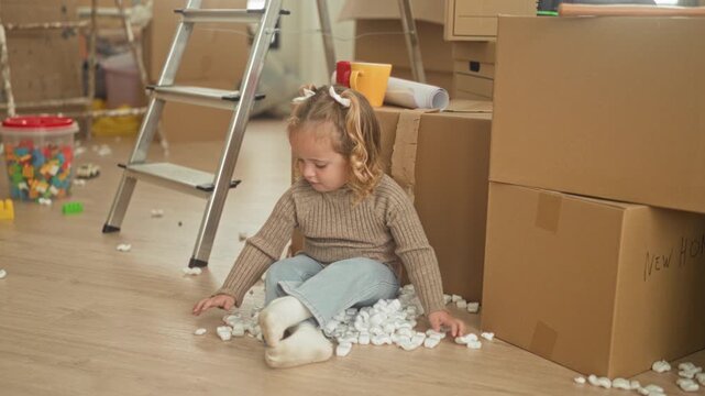 Girl sitting among cardboard boxes and packing peanuts, hand touching foam on the floor in building; quiet curiosity.