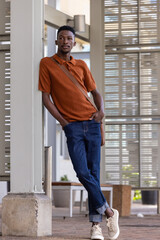Fototapeta premium African man leaning against concrete column at covered walkway, wearing rust polo, sporting tan bag