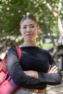 Woman in twenties standing on tree-lined path, carrying pink tote, wearing tan belt and hoops
