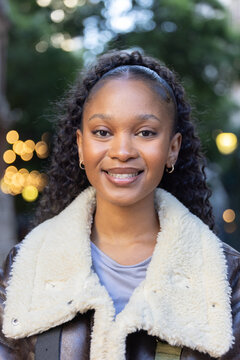 African American adult woman smiling, wearing shearling coat, black headband and gold hoops at park