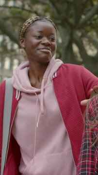 Vertical video: Approaching woman in pink hoodie smiling, hugging friend at campus with notebooks