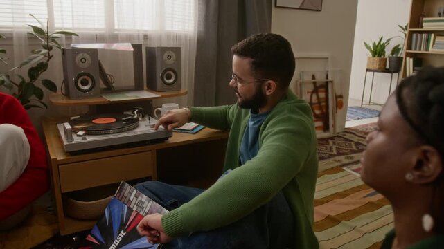 Biracial guy in green cardigan smiling as he setting up vynyl record player while his friends watching, sitting together on cozy living room floor