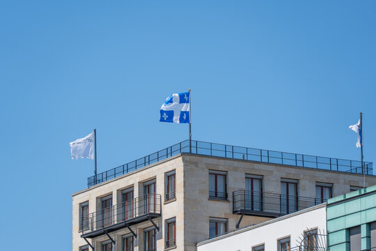 A blue and white Quebec flag with fleur de lis flies over a modern rooftop. Clear sky and pale stone architecture give the city scene a crisp travel mood.