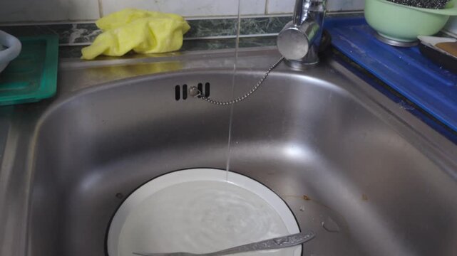 Close-up view of a stainless steel kitchen sink with water running from a chrome faucet. A white plate is submerged in the basin. On the countertop, there are cleaning supplies: a yellow microfiber cl