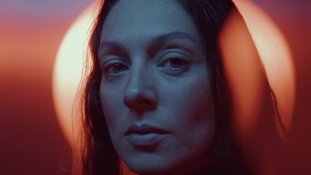 Beautiful woman looking at camera against circular illuminated backdrop glowing with warm orange light. Video portrait