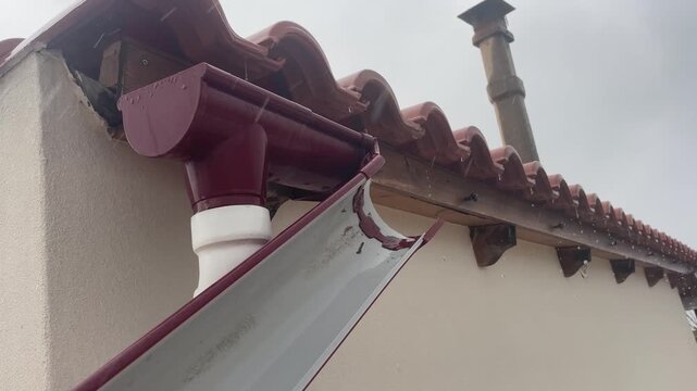 Close up footage of a broken plastic roof gutter system damaged by strong wind with rainwater overflowing and leaking directly onto a wet wooden fascia beam outdoor. High quality 4k footage