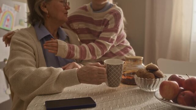 Caucasian girl serving table, putting on tea pot on table and hugging her beloved grandmother