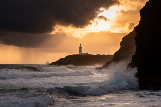 Lighthouse on rocky coastline with crashing waves at dramatic sunset