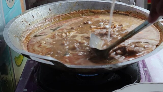 Thick white coconut milk being poured into a large traditional wok filled with simmering Indonesian beef rendang and aromatic spices.