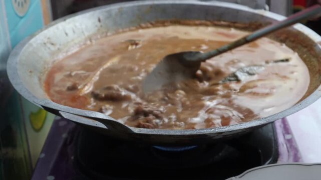 Indonesian beef rendang being stirred and simmered slowly over a gas stove in a large traditional wok with rich spices.