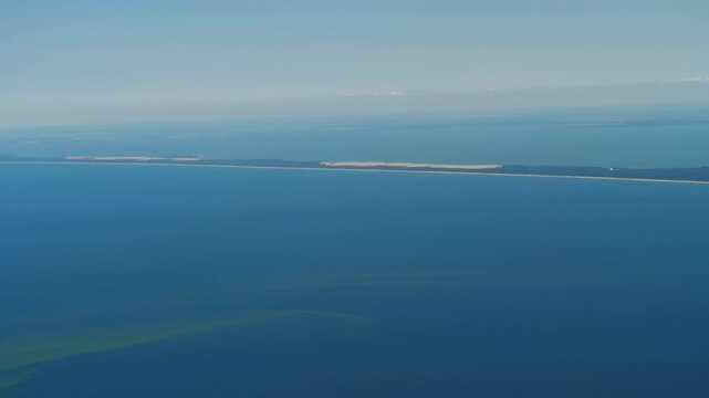 Curonian Spit aerial view dividing Baltic Sea and Curonian Lagoon under a clear blue sky, filmed from a high altitude airplane window shot.
