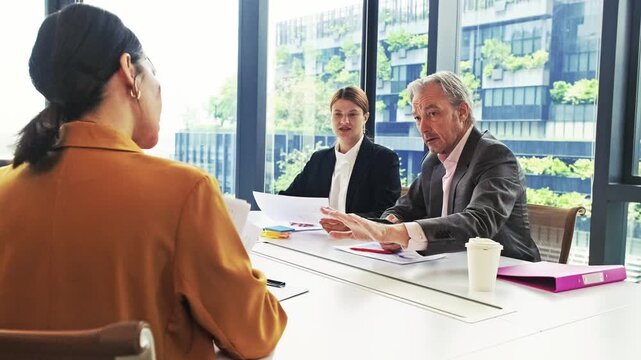 Job interview in a modern office: two interviewers speak with a candidate across a desk, reviewing papers and discussing qualifications with a city view through large windows.