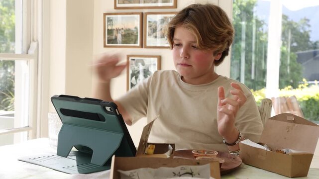12 year old boy eating box lunch in kitchen using tablet computer