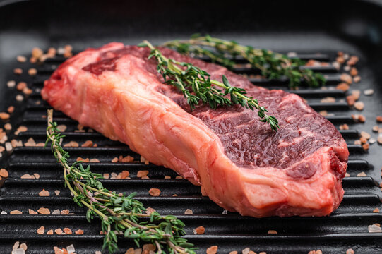 Raw striploin new york strip steak with thyme and salt on a grill pan over a dark stone background, showcasing marbled beef texture and rustic cooking style.