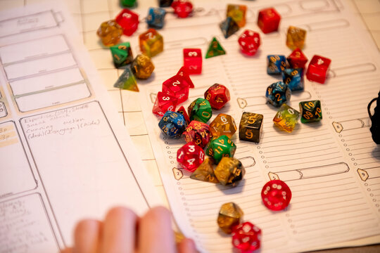 A pile of multicolored polyhedral dice sits scattered across a role playing game character sheet as a hand reaches in. Tabletop gaming in progress on game night.
