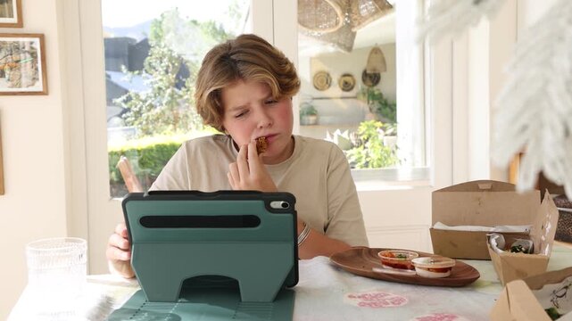 12 year old boy eating box lunch in kitchen using tablet computer