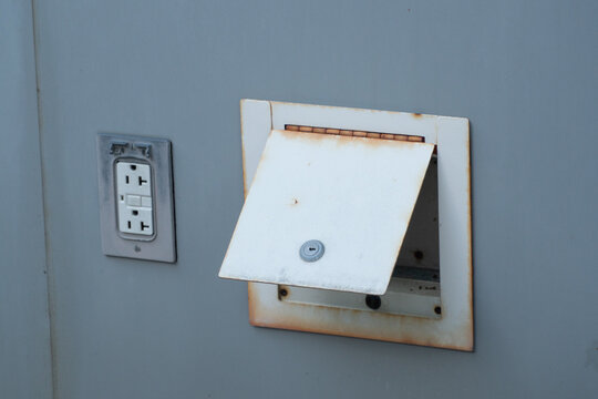 Aged electrical utility box with significant rust and an open access door, revealing a keyhole lock. A worn power outlet is visible on the distressed gray wall.