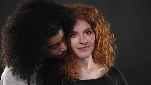 An interracial couple s relationship and connection. A young man with dark curly hair embraces a woman with red curly hair from behind, looking at her affectionately in a studio setting. This footage 