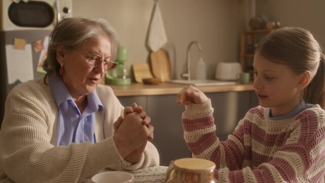 Medium shot of Caucasian granny and granddaughter fooling around, communicating while sitting at dining table in kitchen at home