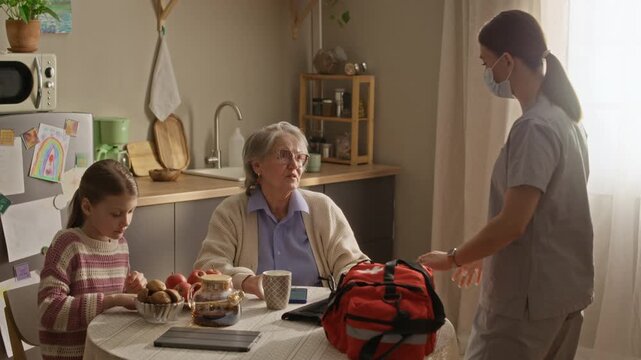 Granny and her granddaughter sitting at table while paramedic greeting her and asking about health