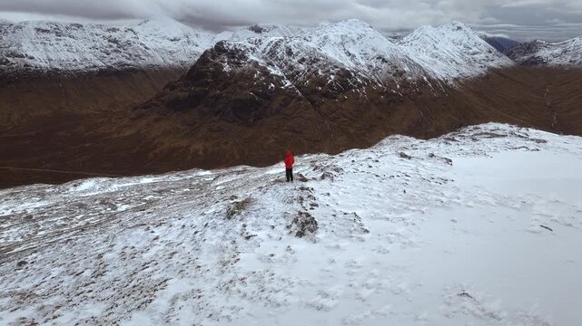 Aerial orbit shot of one male hiker standing on a snowy ridge with rugged mountains in the background.