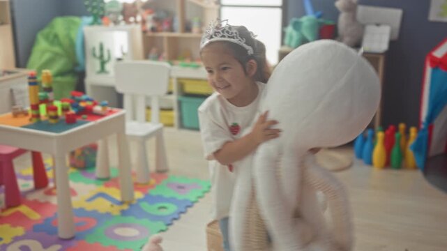 Hispanic girl reaches hand to wooden abacus beside stuffed elephant and red striped play tent, wearing tiara while stepping over scattered toys in building; playful imagination.