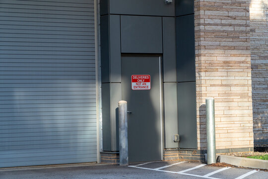 Deliveries Only sign on a grey industrial door next to a large roller shutter and light brick wall, indicating restricted access for service, logistics, and supply chain operations at a commercial.