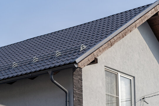 Modern residential house exterior featuring durable dark grey metal tile roof with integrated snow guards and efficient rain gutter system against clear blue sky background.