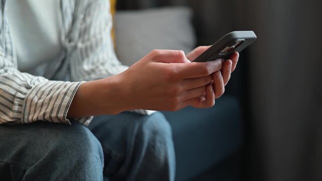 A person sits on a couch, focused on their smartphone while engaging with it. The individual appears to be an adult male, casually dressed, using the device to browse or communicate