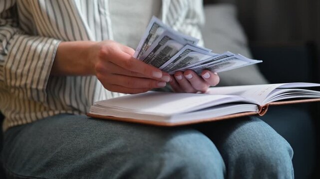 A person sorts and counts cash while sitting on a sofa with a notebook open on their lap. The individual is focused on organizing the bills, creating an atmosphere of financial planning