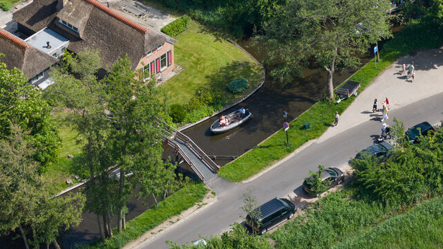 Aerial view of a boat navigating a narrow canal under a wooden bridge near traditional thatched-roof houses and lush gardens in Giethoorn, Overijssel, Netherlands.