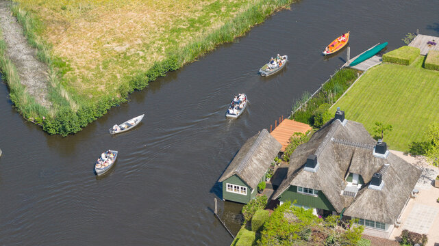 Aerial view of tourists in small boats navigating the narrow canals past traditional thatched-roof houses and lush green gardens in Giethoorn, Overijssel, Netherlands.