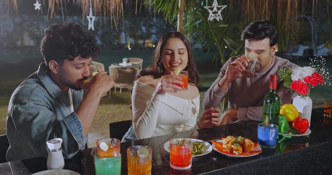 Indian young adult gen z relax male female group sitting around bar table talk speak have fun joy picnic trip. happy girl boy man pick up glass do cheers drink juice spend good time enjoy night place