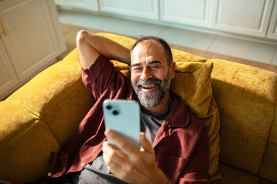 Smiling bearded man using smartphone at home on couch