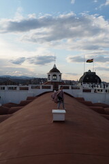 Naklejka premium Young woman tourist standing on rooftop of colonial building in Sucre Bolivia enjoying scenic travel destination with historic architecture terracotta tiles and national flag at sunset