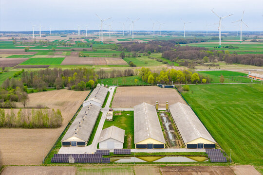 Aerial view of a large agricultural farm with industrial buildings, solar panels, and a wind farm with turbines in the background in Zuromin, Masovian Voivodeship, Poland.