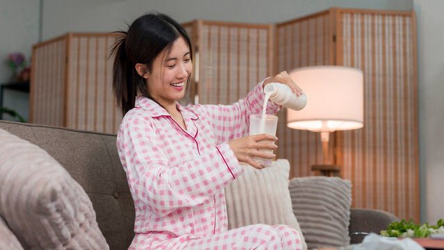 young Asian women pouring milk into glass morning routine breakfast drink healthy lifestyle wearing pink pajamas sitting on sofa couch relaxing at home living