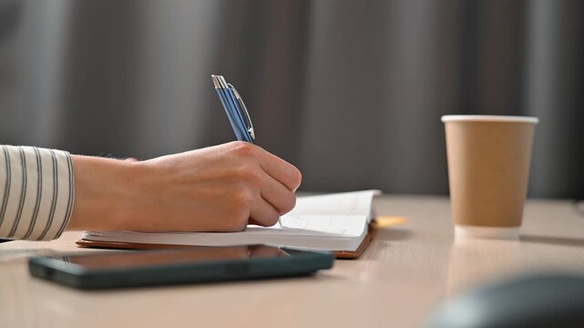 A person is seen turning a page in a notebook and writing notes at a table. The setting includes a coffee cup, a pen, and a mobile device, suggesting a study or work environment