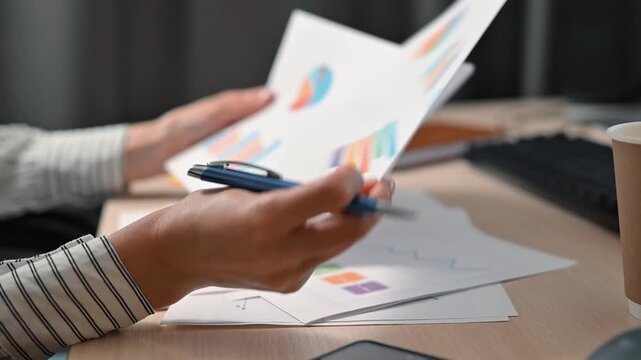 A person is working at a desk, reviewing documents with various charts and graphs. The individual uses a pen to take notes while focusing on the papers in front of them