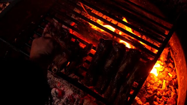 Unidentifiable person carefully turning succulent pork ribs on a barbecue grill over the bright orange glowing embers of a campfire, slowly cooking the meat during a peaceful night outdoors