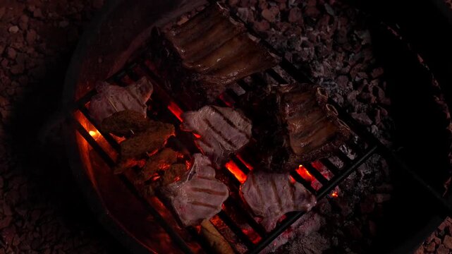 Delicious pork chops and ribs sizzling on a grill over hot embers in a fire pit during a nighttime barbecue, with a hand turning the meat to cook it perfectly over the open flame