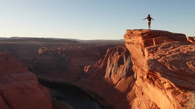 Young adventurous woman standing on the edge of a cliff at horseshoe bend, spreading her arms wide to embrace the spectacular view of the colorado river and red rock canyon at sunset