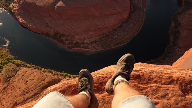 Personal point of view of a man sitting on the edge of the cliff at horseshoe bend, dangling his legs over the famous canyon and the colorado river on a beautiful sunny day in arizona