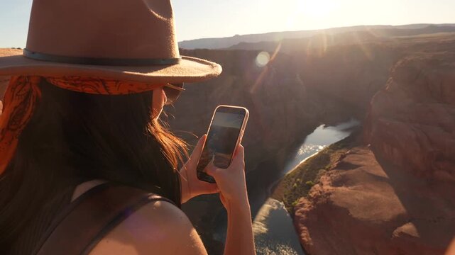 Young female traveler with a hat standing on the edge of the canyon, using her smartphone to capture the stunning scenery of horseshoe bend and the colorado river in arizona during sunset