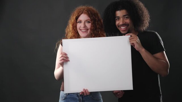 A diverse couple is holding a blank white sign, offering ample copy space for custom messages or advertisements. Both individuals are smiling warmly and looking directly at the camera, conveying a pos