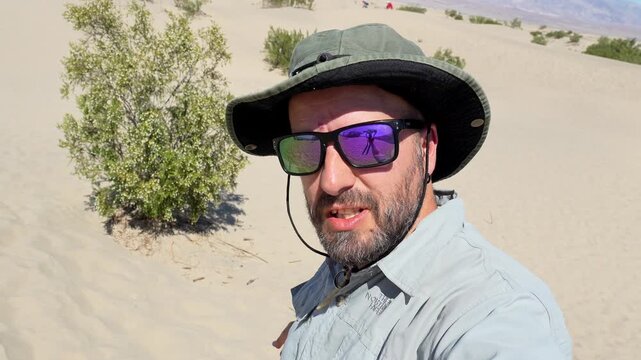 Young male content creator taking a selfie video and talking to the camera while walking in the mesquite flat sand dunes of death valley national park, california, on a sunny summer day