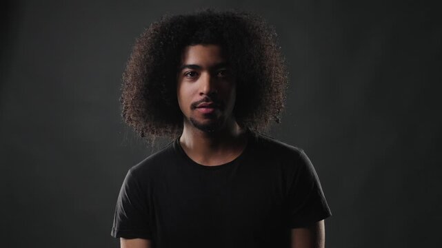 A thoughtful young African American man with voluminous curly hair and a simple black t shirt is captured in a studio portrait. He appears to be pondering deeply, with a serious and contemplative expr
