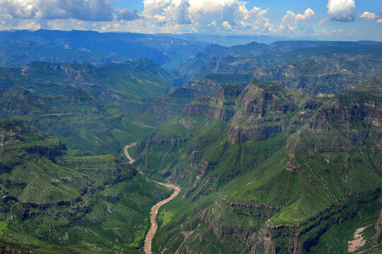 Aerial view of Barrancas del Cobre with a winding river flowing through deep green canyons under a cloudy sky in Chihuahua, Chihuahua, Mexico.
