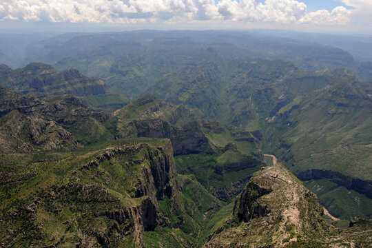 Aerial view of Barrancas del Cobre showing the vast green canyon system with steep rocky cliffs and a winding river under a cloudy sky in Chihuahua, Chihuahua, Mexico.