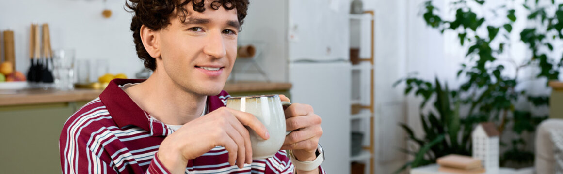 Curly young man enjoying coffee in his stylish modern apartment during leisure time
